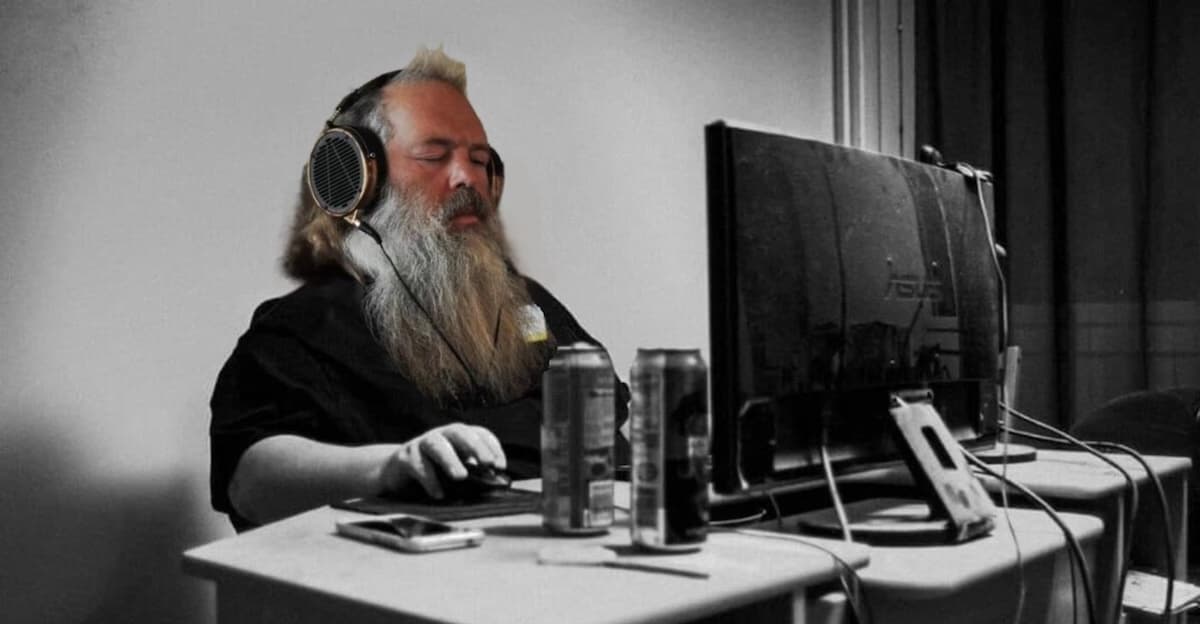 A bearded man in headphones, eyes shut, hunched at a monitor between two energy drinks
